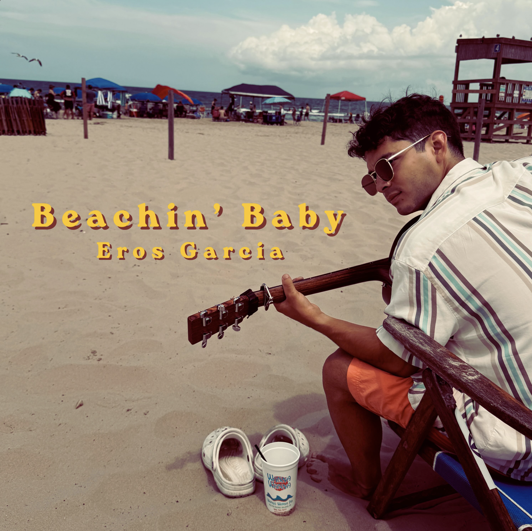 Eros Garcia seated on a beach with an acoustic guitar for Beachin’ Baby